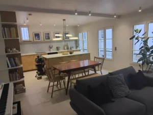 Pièce de vie lumineuse avec canapé gris au premier plan, table de repas en bois, cuisine ouverte et grandes fenêtres.
