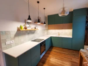 Grande cuisine d'angle sur mesure avec façades vert canard et bois clair, plan de travail blanc immaculé, crédence en zelliges vert d'eau, suspensions noires et parquet en bois massif.