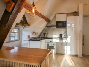 Cuisine mansardée aménagée avec meubles blancs, crédence effet marbre, four encastré et poutres en bois brut.