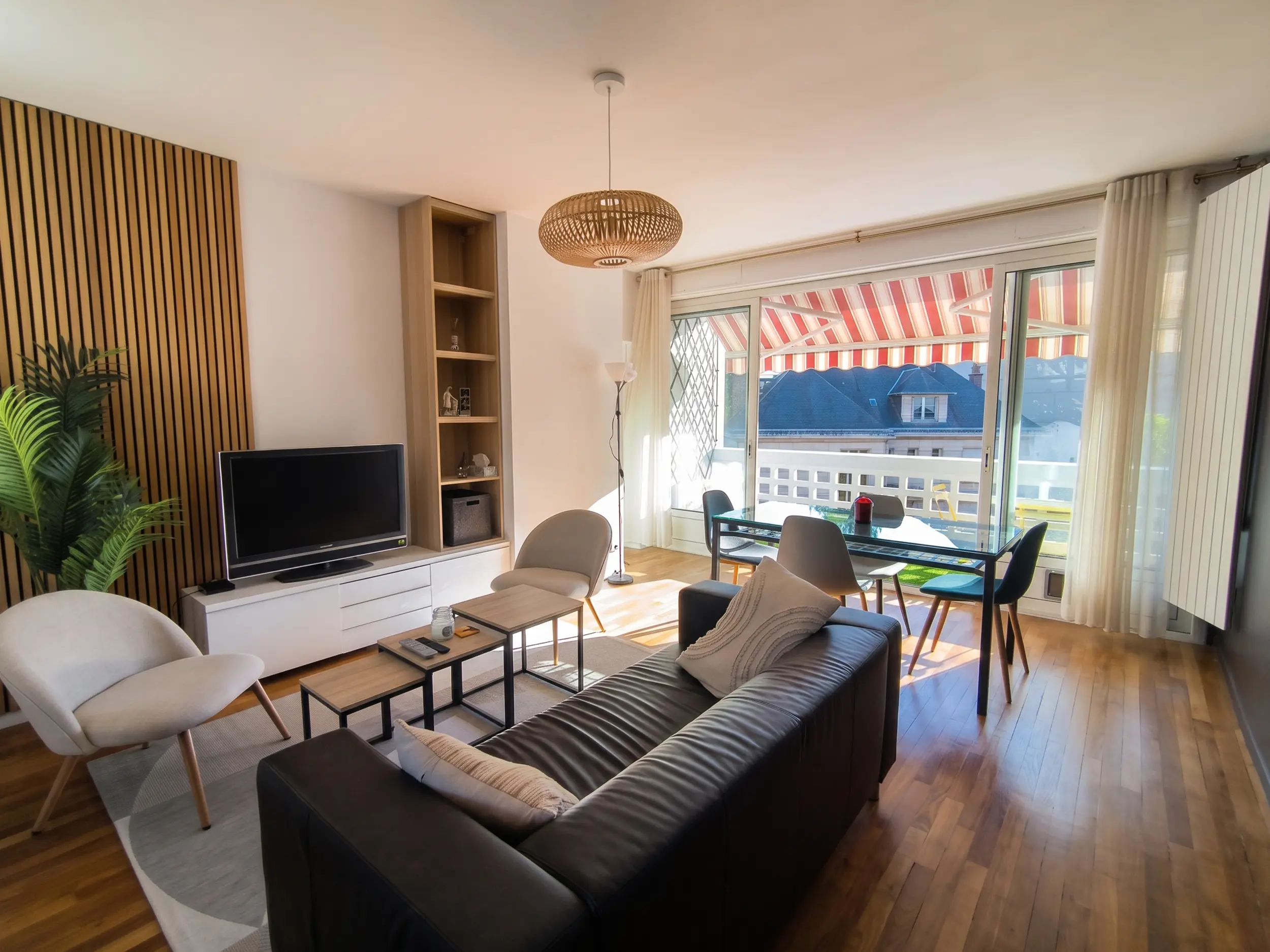 Vue d'ensemble du séjour lumineux avec canapé en cuir noir, fauteuils beiges, table à manger en verre, suspension tressée et grande baie vitrée ouvrant sur le balcon.