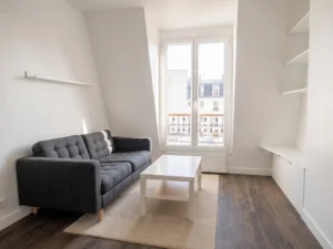 Vue d'ensemble du séjour mansardé avec canapé capitonné gris anthracite, table basse blanche sur tapis clair, sol imitation parquet foncé et menuiseries sur mesure.
