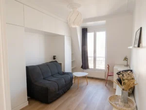 Pièce de vie principale avec canapé convertible gris anthracite intégré sous un pont de meuble sur mesure blanc toute hauteur, espace bureau, table basse tripode et porte-fenêtre lumineuse.