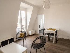 Vue d'ensemble de la pièce de vie mansardée associant coin repas, fauteuils ronds en fil noir, rangements bas et porte-fenêtre lumineuse ouvrant sur les toits.