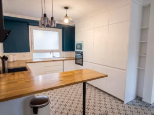 Cuisine ouverte rénovée avec meubles blancs sans poignées, plan de travail et bar en bois clair, sol en carreaux de ciment à motifs et mur d'accent bleu roi autour de la fenêtre.