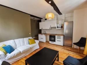 Pièce de vie avec canapé blanc aux coussins colorés, table basse noire, mur d'accent vert olive, cuisine ouverte blanche avec grand réfrigérateur inox et parquet en bois massif.